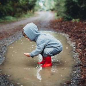 A little boy in red galoshes squats in a muddy water puddle